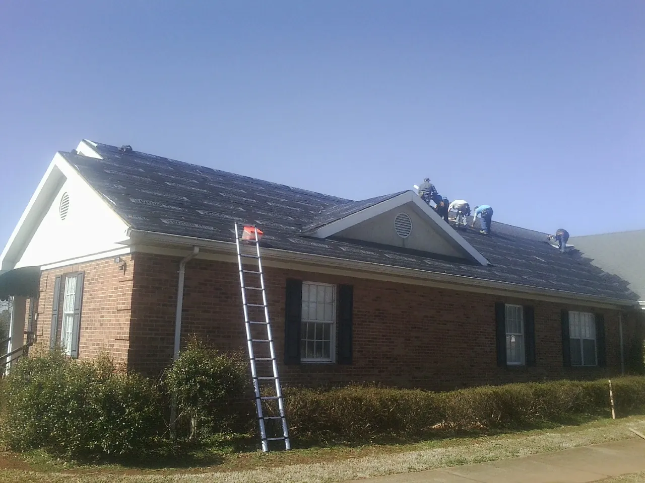 Crew tearing shingles off a brick ranch roof with a ladder set — Metal Roof Repair work by our crew in Pembroke